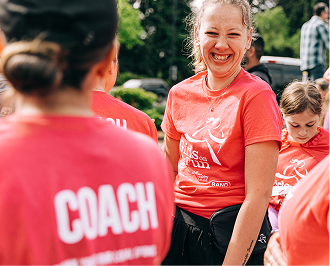 A smiling female coach in a pink t-shirt with the word "COACH" on the back looks over her shoulder. Another coach and a young girl are visible in the background.